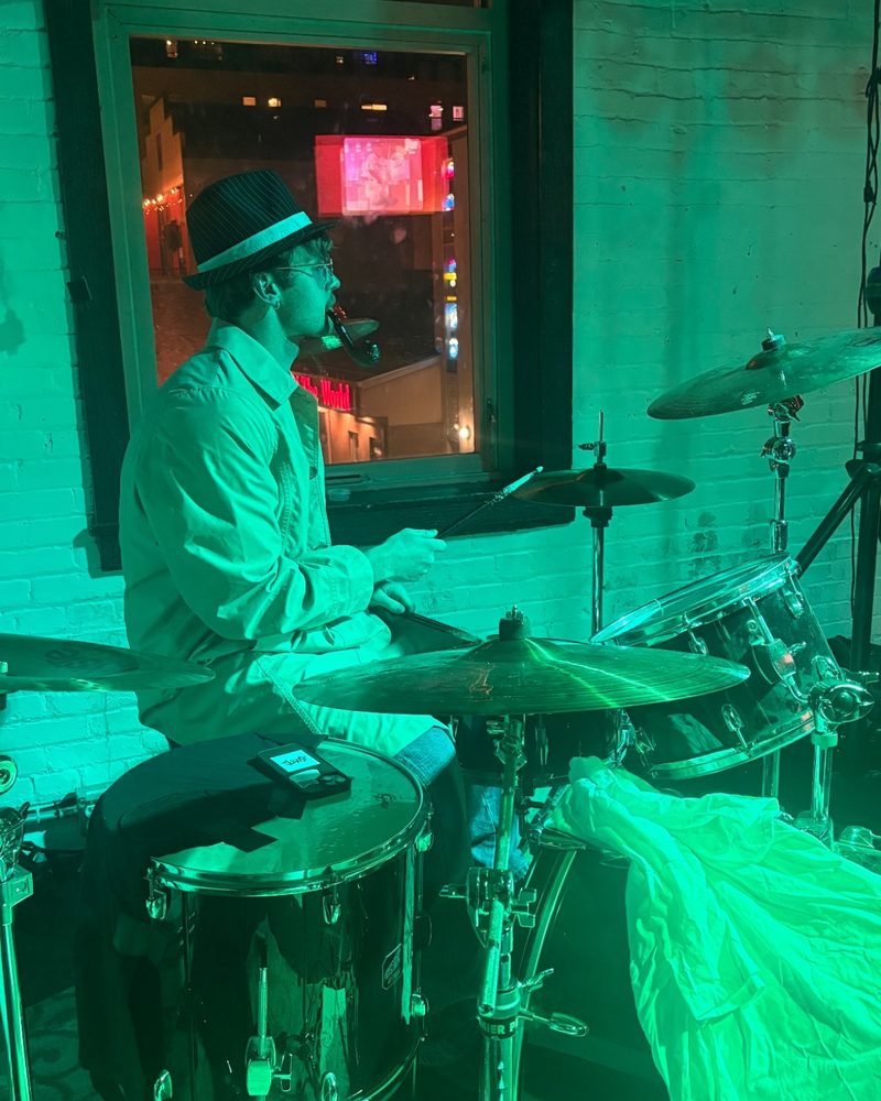 The drummer at his kit under green stage light, Halloween 2025